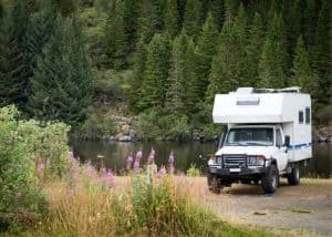 This image displays a white camper van parked by a calm lake with dense pine trees, ideal for outdoor adventure and travel enthusiasts.